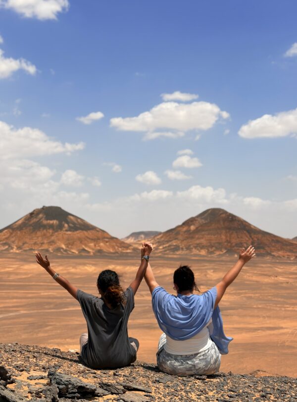 Two travelers with arms raised on a rocky ridge facing the Black Desert hills