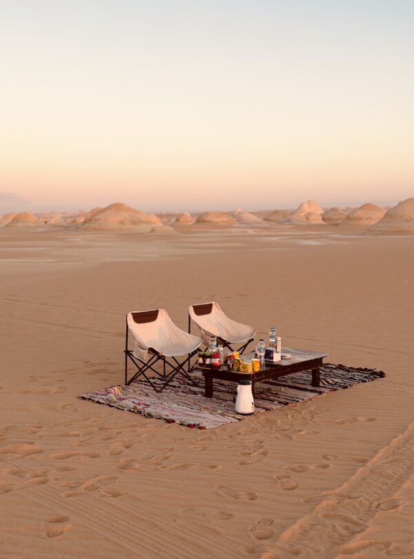 Two camping chairs and a low table set on a rug in the White Desert at sunset